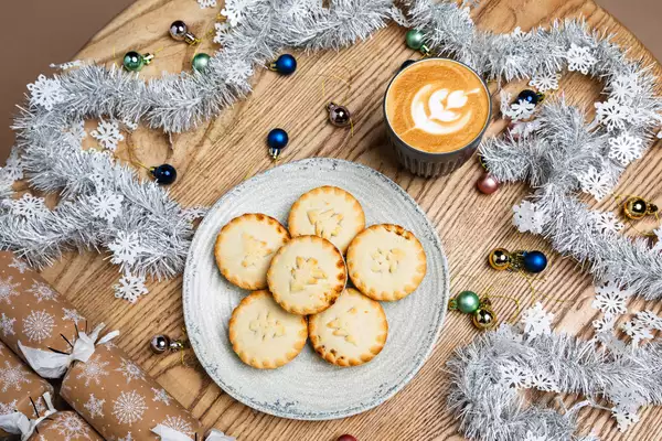 A festive table with six mince pies on a plate, a latte with leaf art, silver tinsel, snowflake cutouts, colorful baubles, and gift crackers.