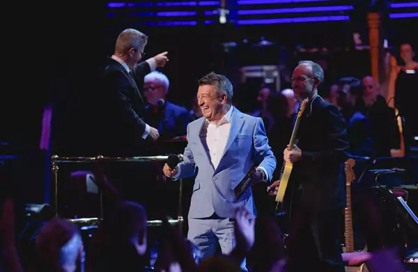 A singer performing in a grey suit with a full orchestra at the BBC Proms