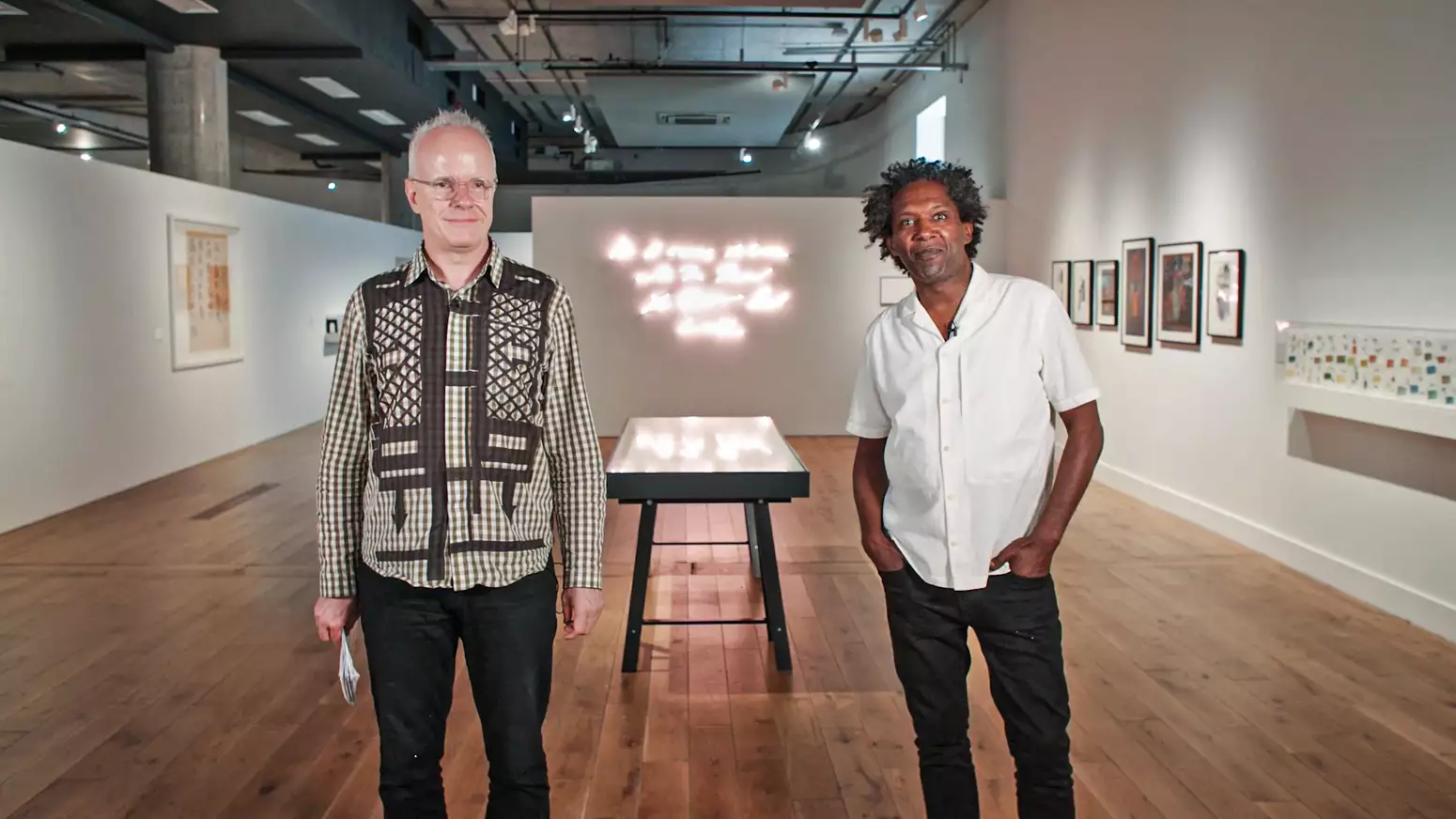 Hans Ulrich Obrist and Lemn Sissay stand in the Poet Slash Artist exhibition at HOME during MIF21. Hans Ulrich stands on the left; he is white with a pale peach skin tone and very short white hair; he wears glasses with clear frames, a green check shirt with black lattice and arrow patterns and dark navy jeans; he is smiling and looking just to the right of the camera. Lemn stands on the right; he is Black with a deep brown skin tone and medium length black and grey afro hair; he wears a white short sleeve shirt and black jeans, his hands are in the front pockets of his jeans; he smiles with a closed mouth directly at the camera. A vitrine can be seen between them in the background, and beyond that a neon artwork on the wall; a large frame hangs on the left wall behind Hans Ulrich; many smaller frames hang on the right wall behind Lemn. The industrial style ceiling of the space can be seen above the white gallery walls and partitions.