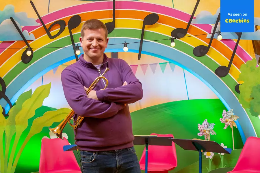 man in a purple sweater holds a trumpet and smiles with his arms crossed in front of a colorful, musical-themed background. The setting includes a large rainbow with oversized black musical notes, green plants, and string lights. Pink chairs and music stands are arranged in the foreground. A blue label in the top right corner reads 'As seen on CBeebies.