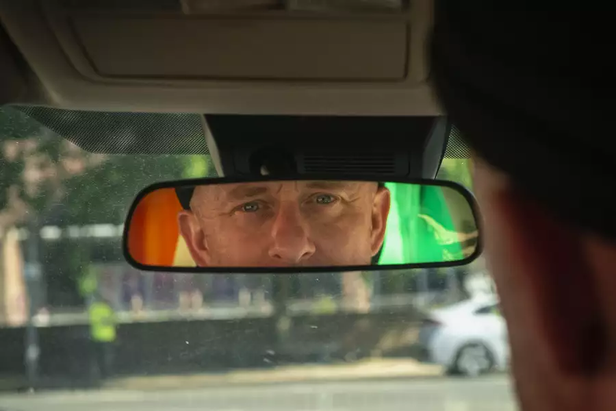 Close-up view of a man's face reflected in a car's rearview mirror. The background shows a blurred outdoor scene with trees and a parked car, while the man's intense gaze is highlighted in the mirror.
