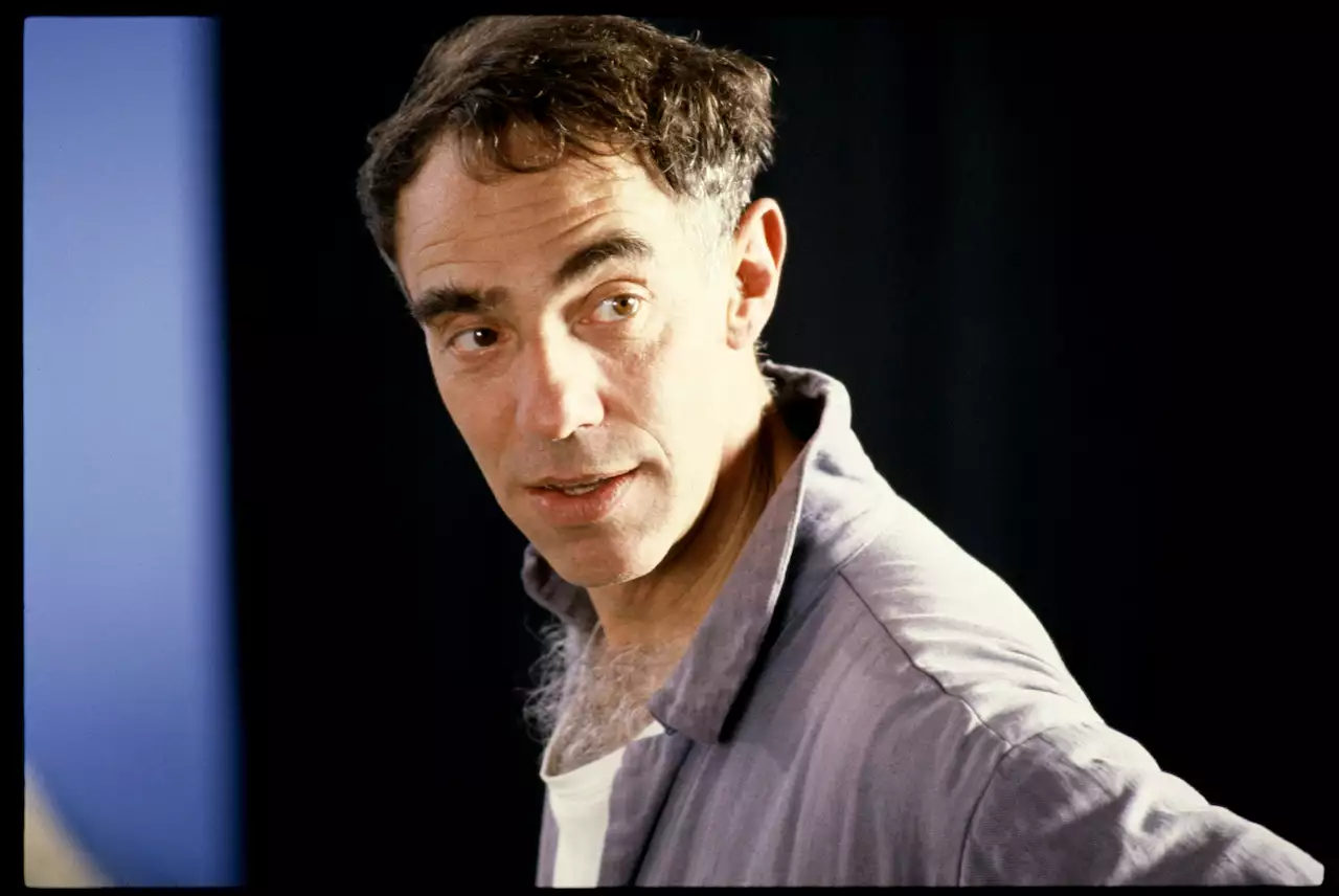 Derek Jarman wearing a light grey shirt and looking off camera. The background is a blurry blue and black.