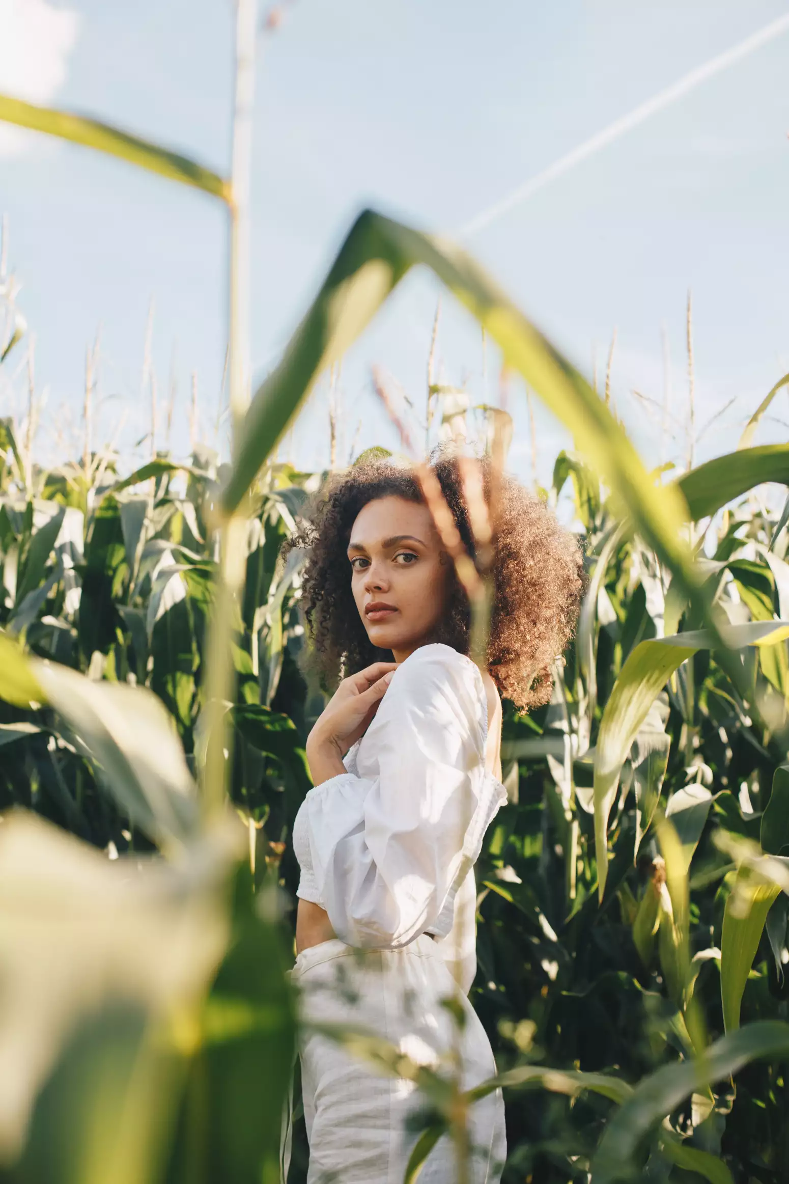 Mali Hayes is stood in a field of leaves that is taller than she is. She's wearing a white top and trouser, stood side on, but looking directly into the camera.