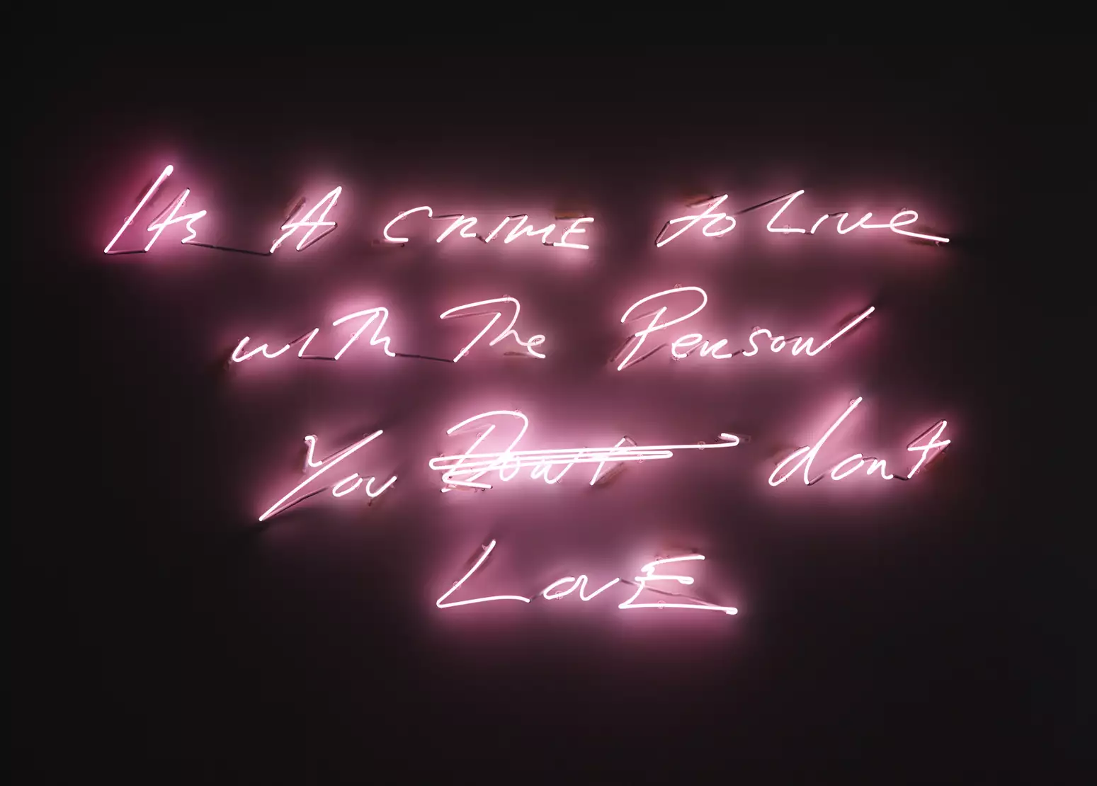 Handwriting rendered in pink neon tubing glows against the wall it is mounted on; the room is in darkness. The text reads: ‘Its A CRIME to Live with The Person You D̶o̶n̶t̶ dont LovE’ [sic]
