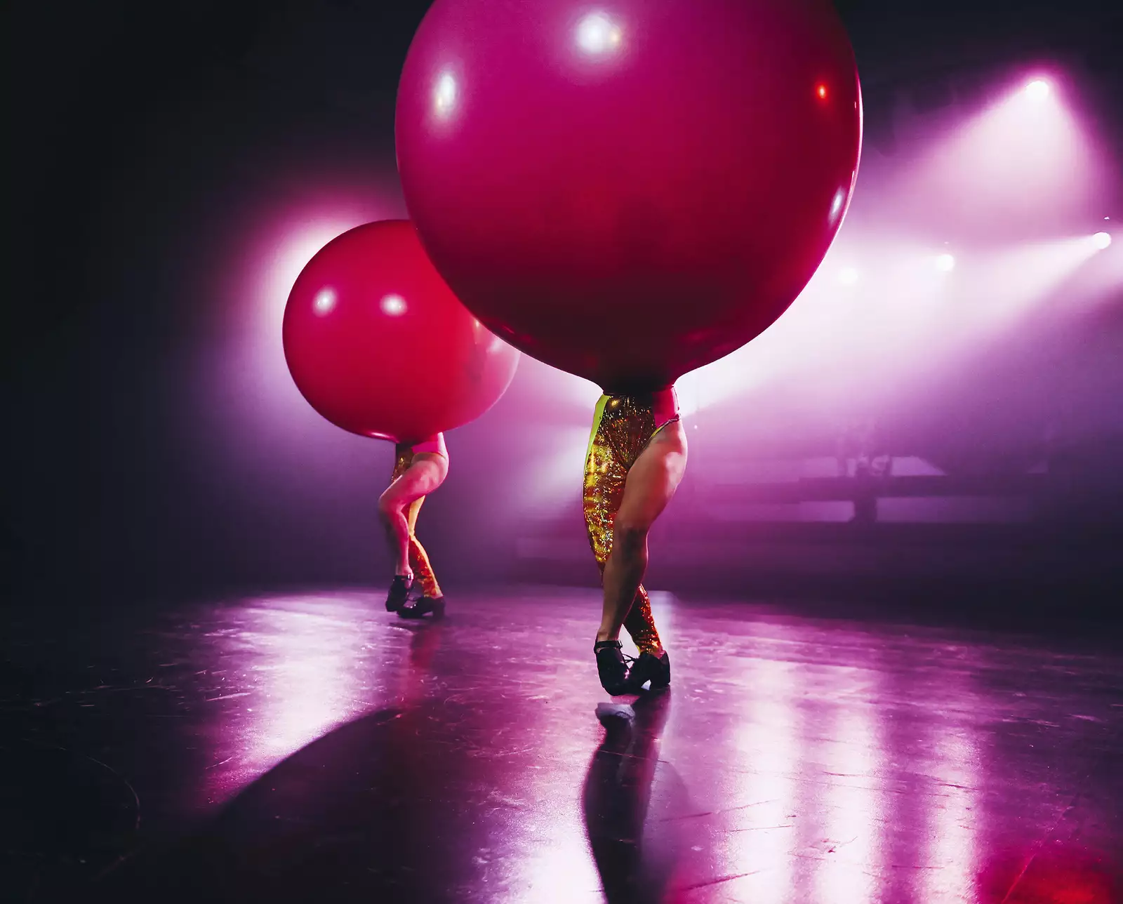 Performers balance large red balloons on their heads while wearing sparkly gold outfits on a brightly lit stage during WAKE.