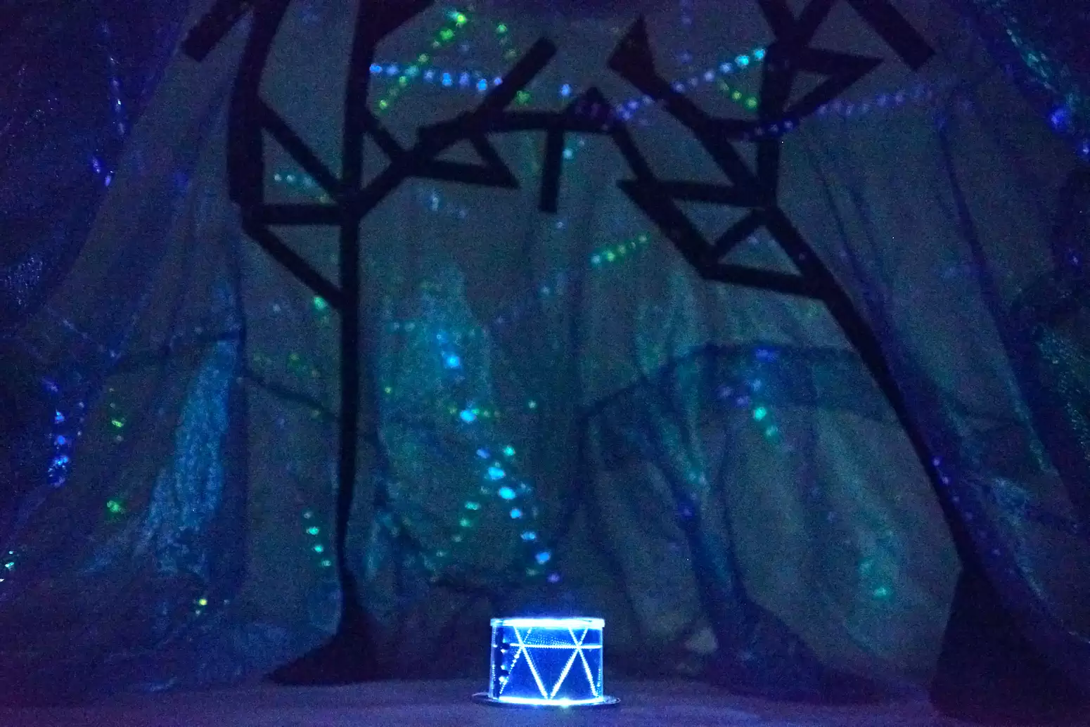 An image taken from inside a planetarium tent, which is made of gauzy blue fabric with black lines marking star constellations on it. A box on the ground glows blue and has patterns where light escapes, projecting green and blue dots of light onto the tent.