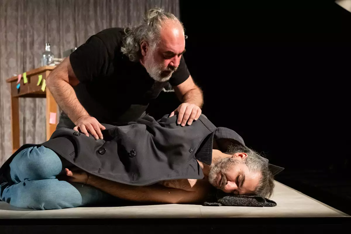A person lying down on stage with eyes closed underneath a coat – with another person peering over him