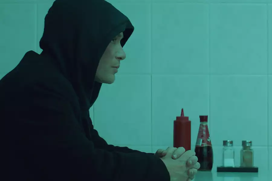 The image shows a person sitting at a table in what appears to be a dimly lit, minimalist diner or café. The person is wearing a dark hoodie with the hood up, facing left, and sitting with their hands clasped in front of them, creating a contemplative or introspective mood. On the table in front of them, there are bottles of ketchup, vinegar, and salt and pepper shakers, adding to the simplicity of the setting. The greenish-blue tint of the lighting adds a moody, cinematic atmosphere to the scene.