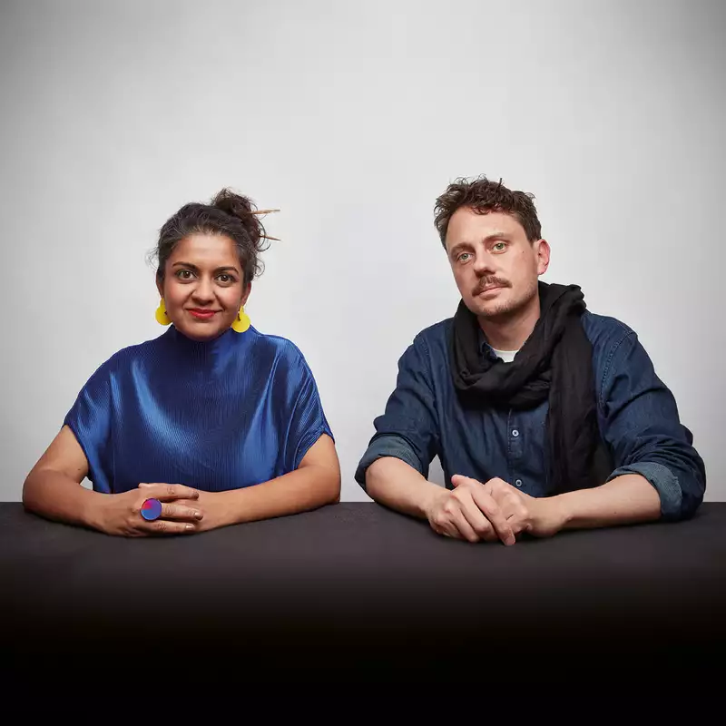 Two members of Superflux sat at a table, looking at the camera. The person on the left is wearing a shiny blue top and yellow earrings. The person on the right is wearing a blue shirt and black scarf.