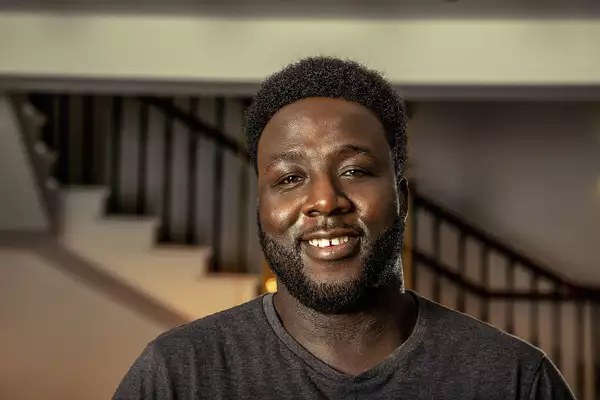 A headshot of a man looking straight at the camera. His shoulders are visible and he wears a dark grey marl t-shirt and has a short black afro.