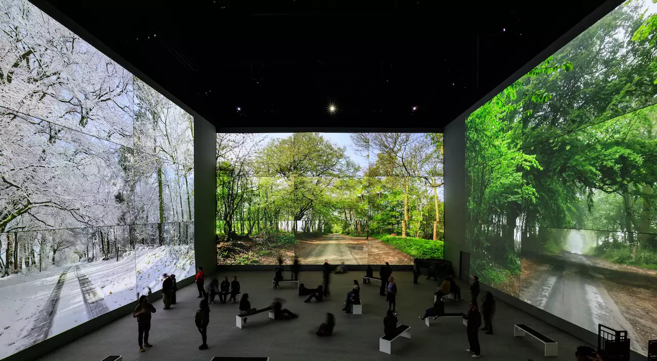 An image from David Hockney: Bigger & Closer (not smaller & further away) showing people sat and stood within the exhibition, surrounded by his artwork showing the changing seasons