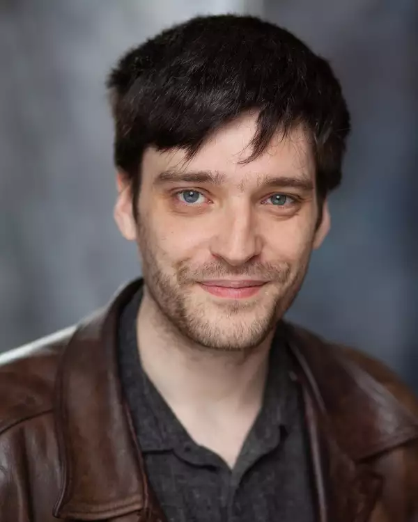 Headshot of Elliot Hughes smiling and wearing a brown jacket