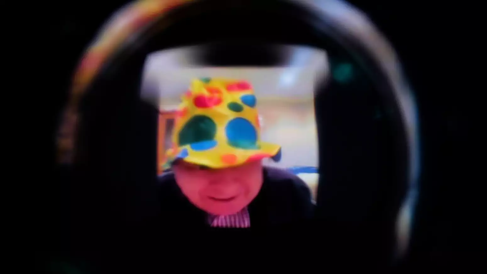 A blurred and light-distorted image down a dark camera lens of an older person with white skin wearing a bright polka-dot clown hat.