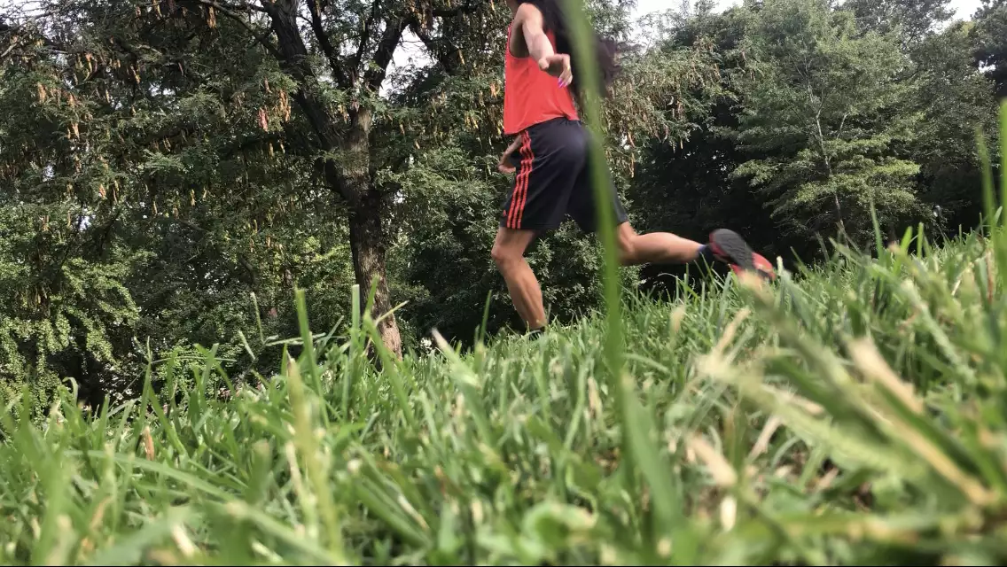A diagonal photo of ​​Benedict Nguyễn, wearing black shorts, orange tank, and black and orange sneakers, lunging forward on left leg facing away from camera with left arm reaching behind her body, in a field with green grass and trees in background.