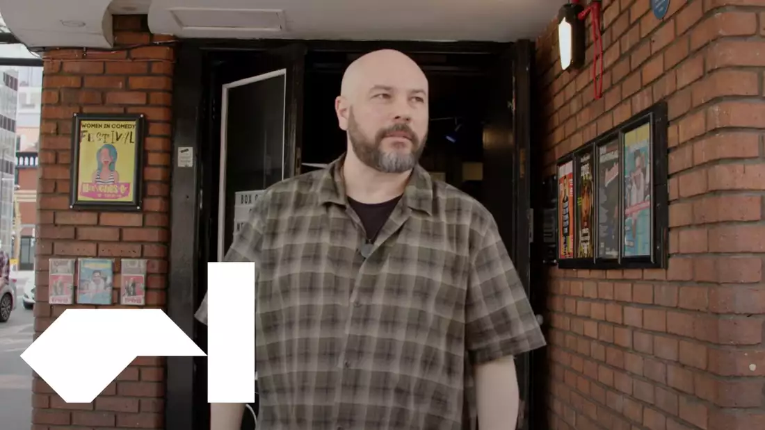 Photograph of a man smiling, looking straight ahead. He has a beard and his shoulders are just visible; he wears a green checked shirt. In the background, advertising posters are visible but out of focus.
