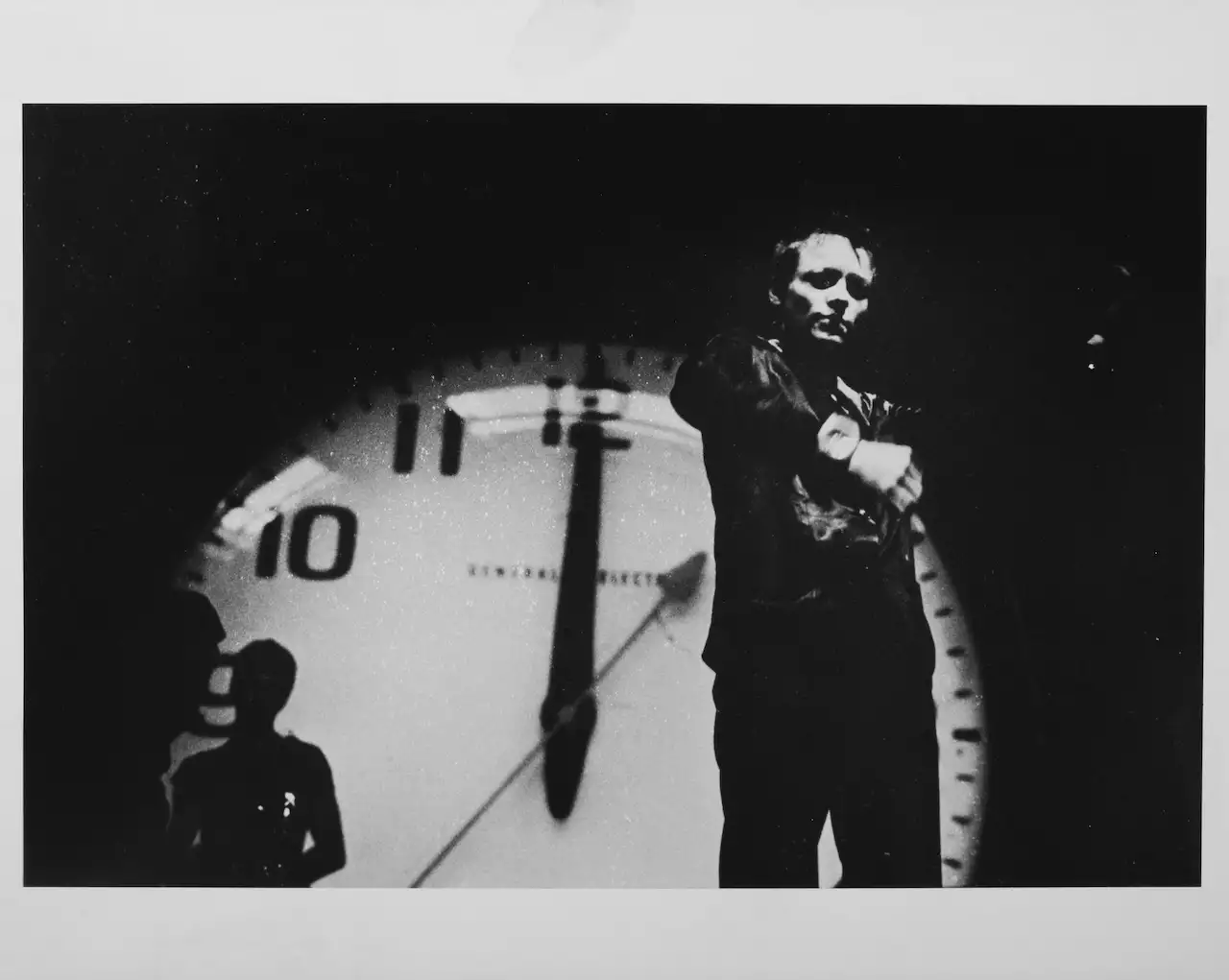 Black and white still from Laurie Anderson's United States Parts 1–4, showing Laurie stood in front of a giant clock