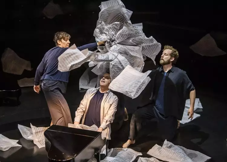 A photograph from Tao of Glass, showing Phelim McDermott sat at a miniature grand piano while two puppeteers construct an oversized wig for him using large pieces of sheet music.