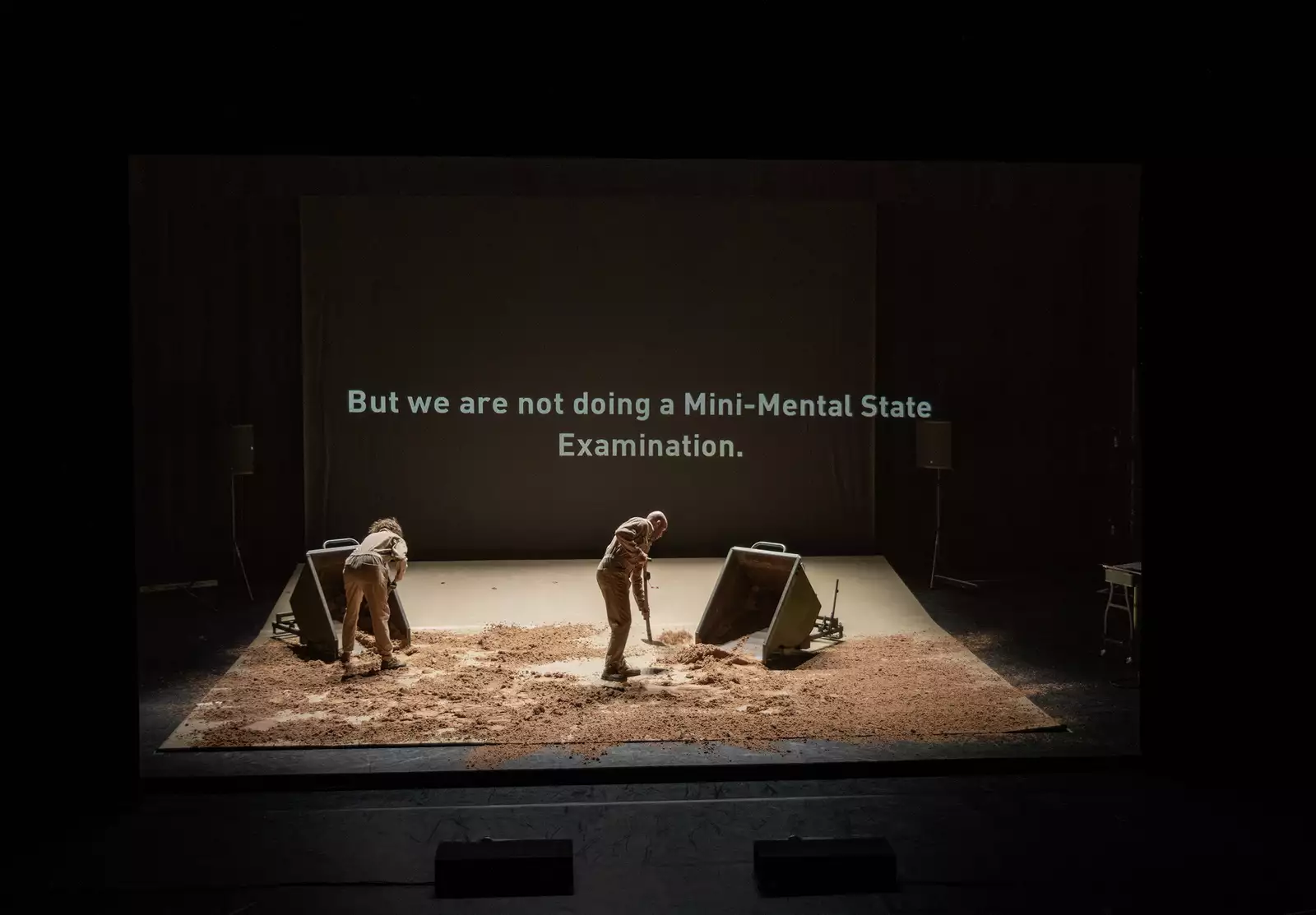 Two people sweeping sand on a stage with the words 'But we are not doing a Mini-Mental State Examination' on a screen behind them