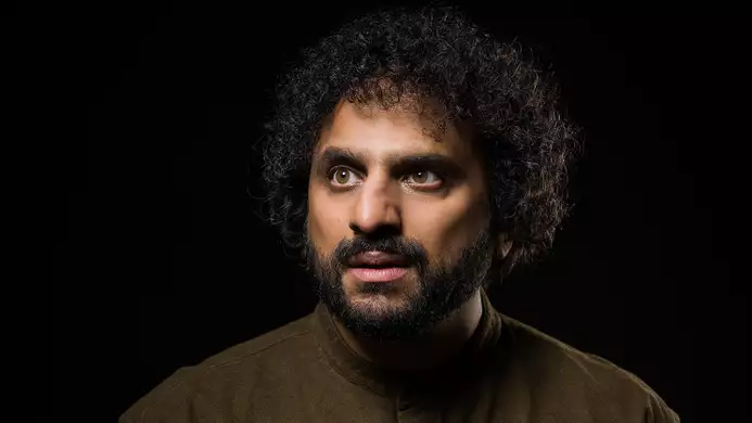 Headshot of Nish Kumar wearing a brown jumper, on a black background