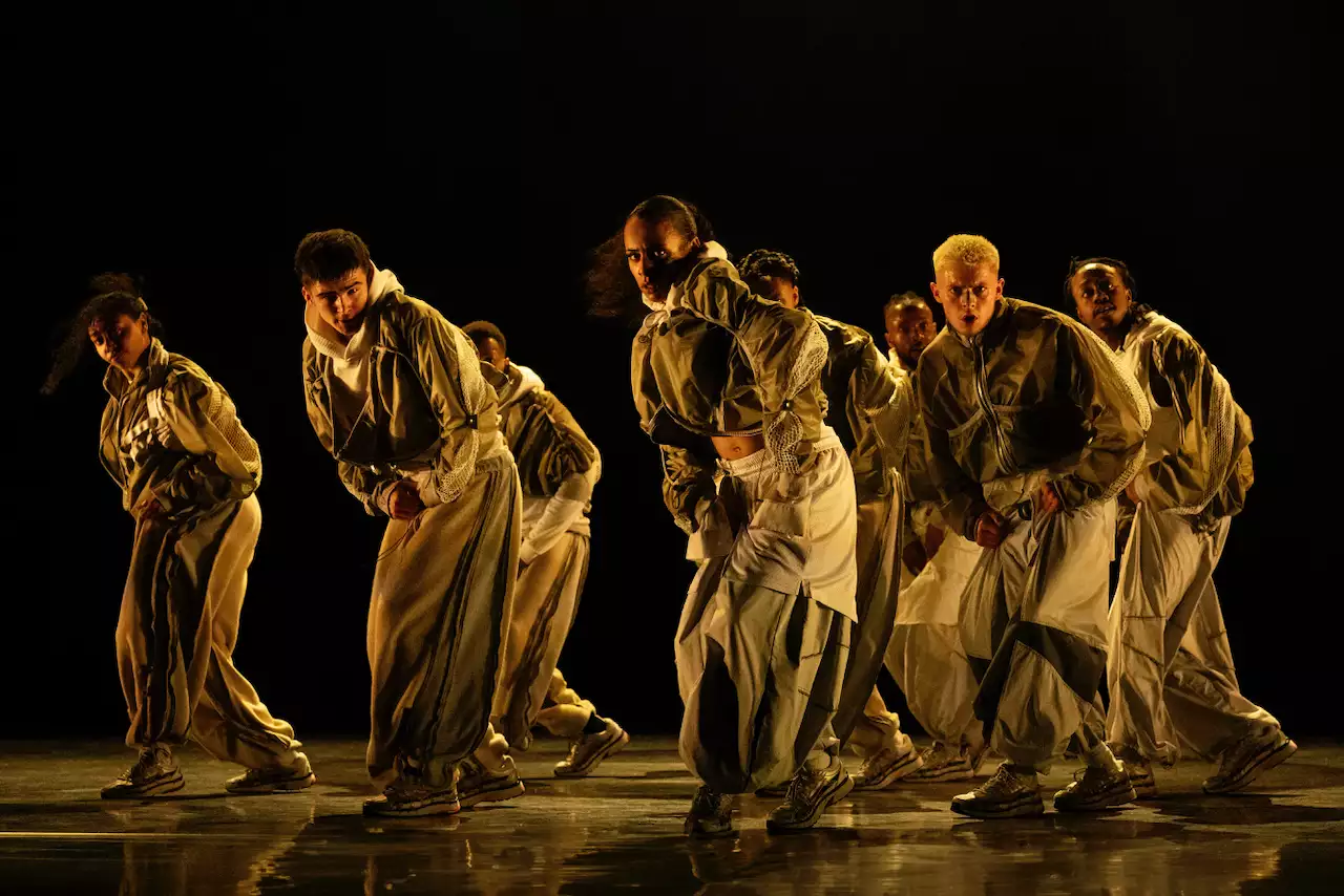 A group of dancers in coordinated white outfits performing under warm, dramatic lighting as part of Cycles.