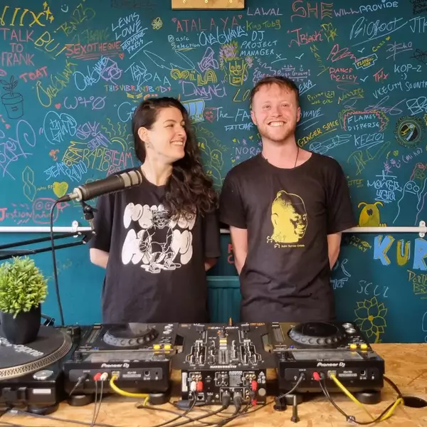 Two members of STEAM radio stood behind recording equipment smiling – in front of a chalk board filled with names