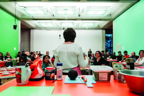 A photograph of a room at Manchester Art Gallery which has been converted into a temporary classroom, with bright green walls on the left and right of the frame. A class of adults are listening intently as the tutor, with her back to us, leads a cookery class.