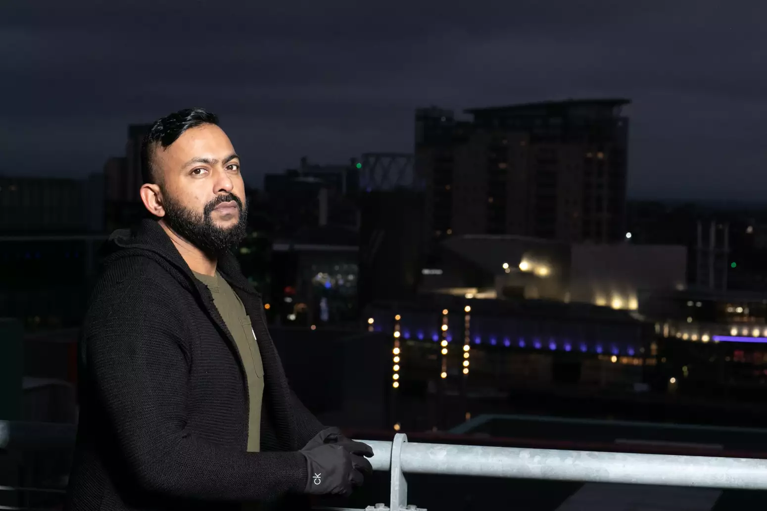 It's nighttime. Sam is stood on a bridge, side on, but looking at the camera. He's wearing a black jacket and gloves. There is a cityscape in the background