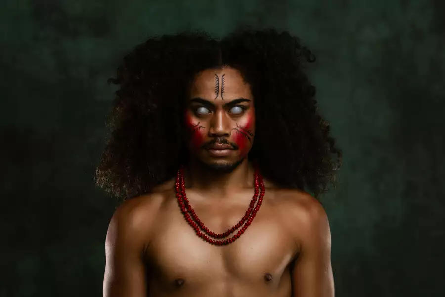 A member of FAFSWAG with glowing white eyes and red and black face paint stares intensely at the camera. They wear red beaded necklaces. The background is dark green.